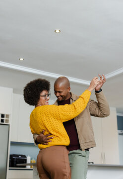 Diverse couple dancing and holding hands high in home kitchen with coffee maker on white countertop