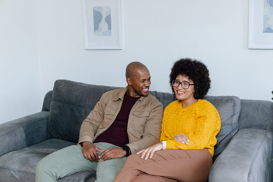 Diverse couple sitting on grey sofa in living room with abstract art prints and potted plant