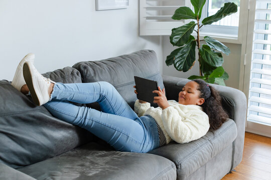 African American child reclining on sofa in living room with tablet, framed print, fiddle fig plant