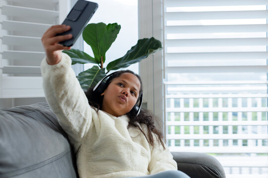 Preteen girl holding smartphone posing for selfie wearing headphones on gray sofa near shutters