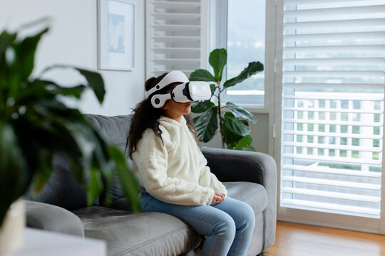 Child wearing VR headset is sitting on gray couch flanked by fiddle fig plants near shutters