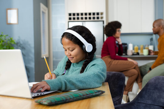 Diverse family in home kitchen with parents holding mugs while girl writing in notebook by laptop