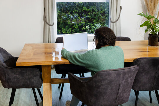 Woman sitting at wooden dining table working on laptop near window, with water glass and plant