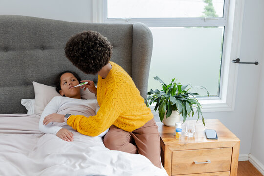 Mother and daughter leaning over bed checking temperature with digital thermometer by bedside table