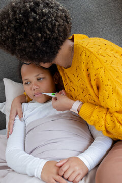 Diverse mother and daughter leaning in bed with mother holding thermometer under child's tongue