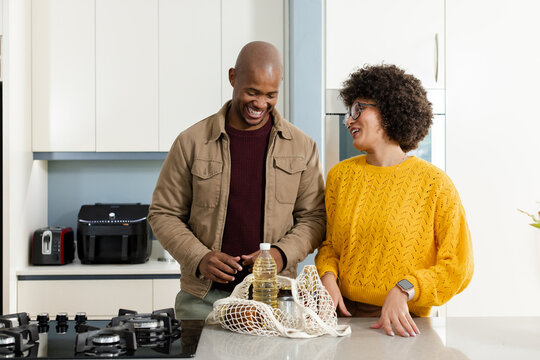 Diverse couple unpacking groceries at marble kitchen island with mesh bag, oil bottle and bread