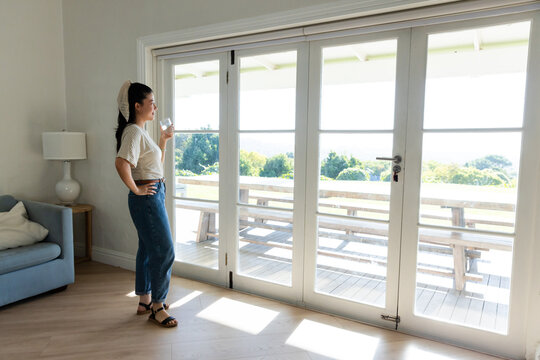 African American woman wearing jeans standing near French doors at home holding glass, copy space