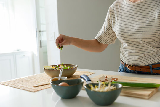 Adult African woman preparing salad, sprinkling herbs at counter with bowl, in shirt, copy space