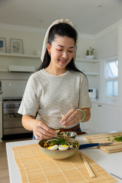 Asian woman standing at kitchen island wearing scarf preparing ramen bowl with halved eggs