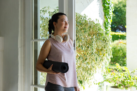 African American woman standing at glass door in pink top, leggings, holding yoga mat and headset