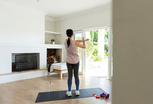 Sunlight urging African American woman in workout clothes stretching on mat at home near dumbbells