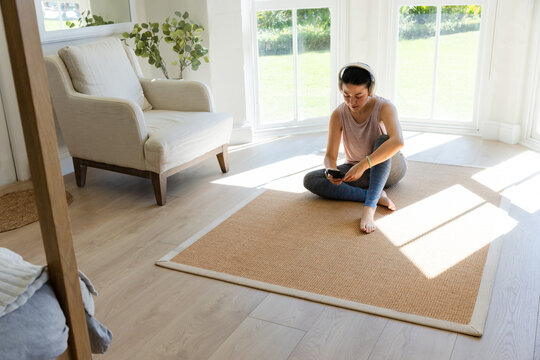 African American woman sitting on rug at home using phone wearing leggings and headset, copy space