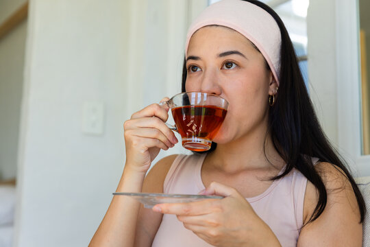 African American woman sipping from clear glass teacup holding saucer at home in pink headband