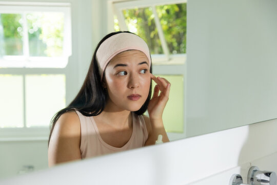 Adult Asian woman wearing headband examining eye, touching temple at bathroom sink with dropper