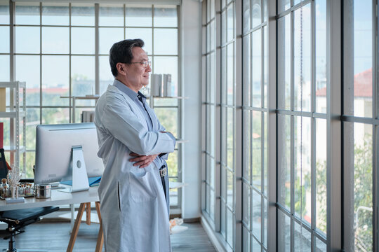 Professional senior Asian CEO doctor resting work, stands and looks out window, motivated to think about medicine research, diagnosis, assessment policy, with duty responsibility and hospital policy.