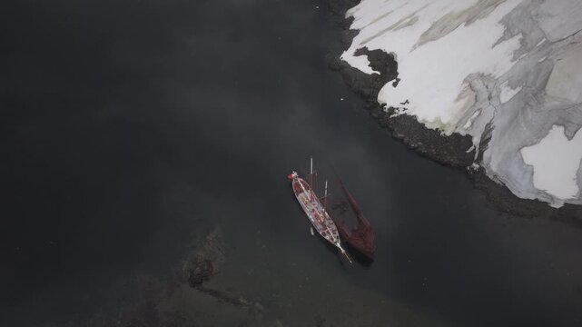A steady bird's-eye view of the schooner Amazone moored to the rusted Governoren shipwreck in Foyn Harbour, Antarctica, as light clouds and subtle sun reflections move across the dark water