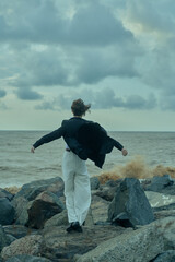 Obraz premium Man stands on a rocky shoreline facing the sea with wind lifting his jacket, calm yet determined mood, coastal landscape, horizon wide, solitary moment of resilience and contemplation amid crashing