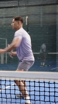 Vertical video: Pivoting man in purple tee swinging paddle, returning ball nearing net on court