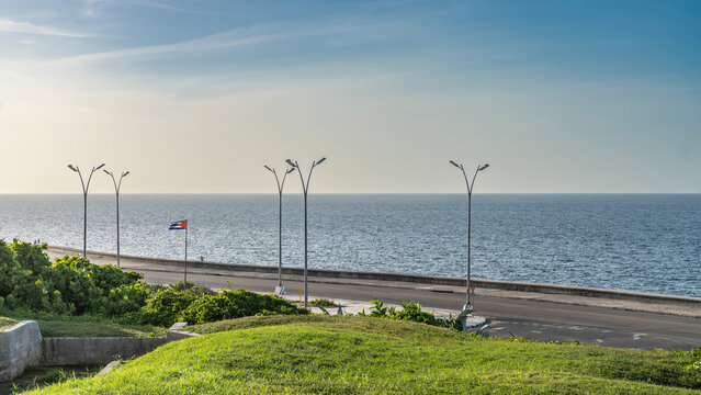 The Malecon Embankment runs along the ocean shore. Street lights along the highway. The flag of Cuba is flying. Green grass on lawns. The blue sky. Havana.