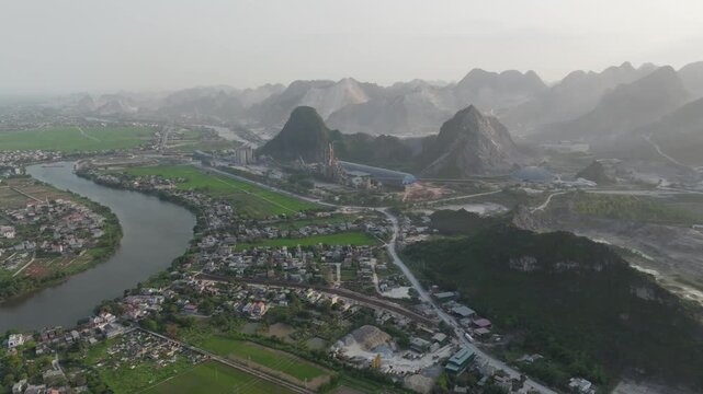 Winding river curves through dense residential areas and green fields, surrounded by dramatic karst formations in Ninh Binh, captured from above at dawn.