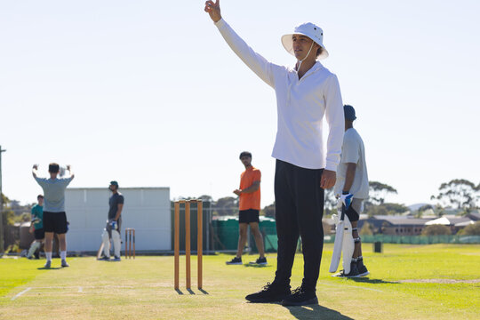 Umpire raising finger and players milling around stumps on grass pitch, under clear sky