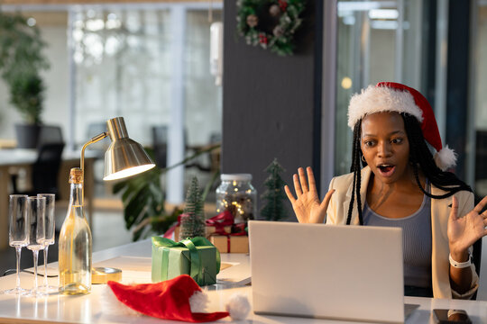 Black woman wearing Santa hat raising hands at office desk with laptop and gifts, copy space