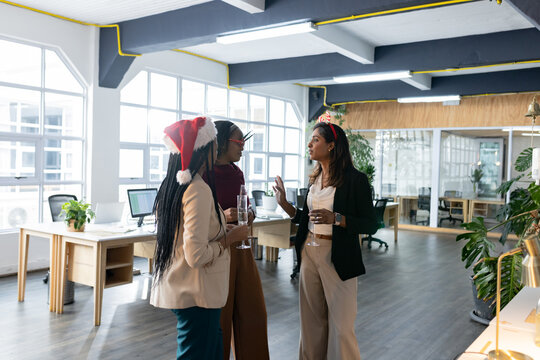 Diverse female colleagues chatting while holding wine glasses in open office with computer monitors