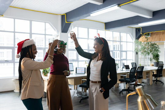 Diverse female coworkers toasting in festive headgear in open-plan office with champagne flutes