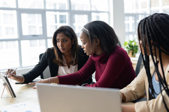 Diverse female coworkers collaborating on tablet with stylus and laptop at office table with charts