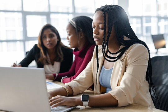 Diverse female colleagues typing on laptop and reviewing printed charts on table in bright office