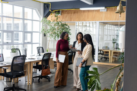 Diverse female colleagues discussing project using tablet laptop and folder in open-plan office
