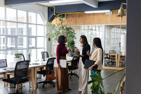 Diverse female coworkers standing in modern office holding white folder portfolio and tablet device