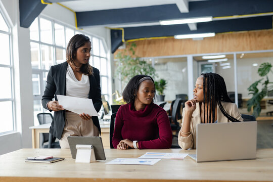Diverse female colleagues studying charts with tablet and typing on laptop at table in open office