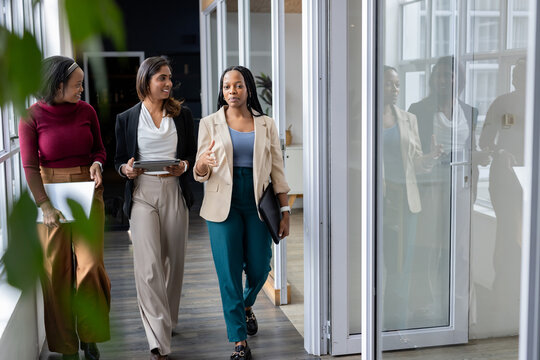 Diverse female colleagues in business attire walking office corridor with laptop, tablet and folder