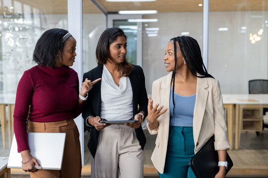 Diverse female colleagues walking through office corridor talking holding laptop tablet smartwatch