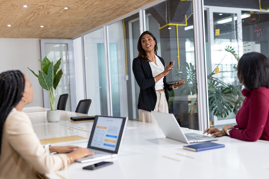 Diverse female coworkers presenting plan on glass wall in meeting room with laptops and marker