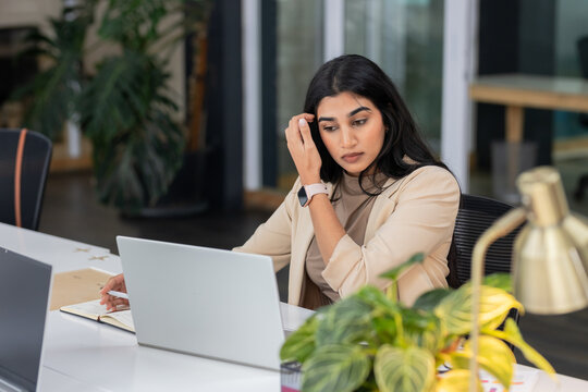 Indian woman wearing blazer typing on laptop and holding pen above notebook in open-plan workspace