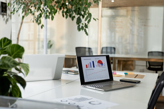 Silver laptop displaying colorful charts, resting next to smartphone on white desk in modern office