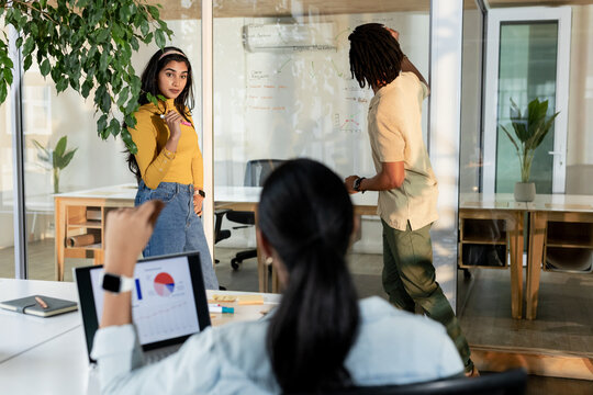 Diverse coworkers collaborating on glass partition whiteboard and laptop charts at conference room