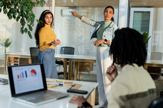 Diverse coworkers collaborating on glass whiteboard in office with laptop charts, copy space