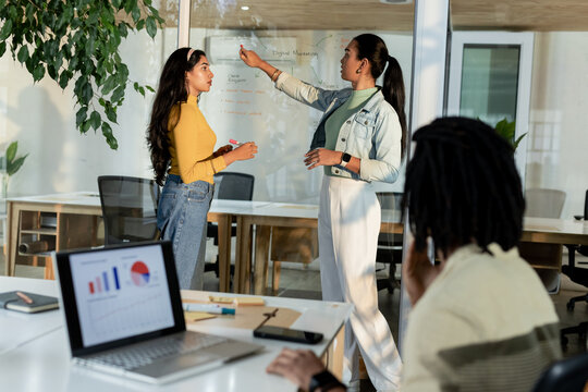 Diverse coworkers discussing digital marketing on glass whiteboard with laptop in conference room