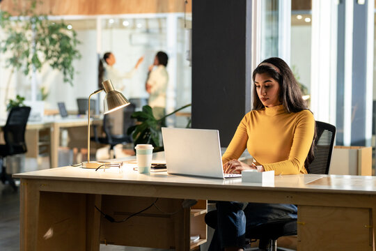 Diverse female coworkers working together at office desk with laptop, lamp, coffee cup, copy space