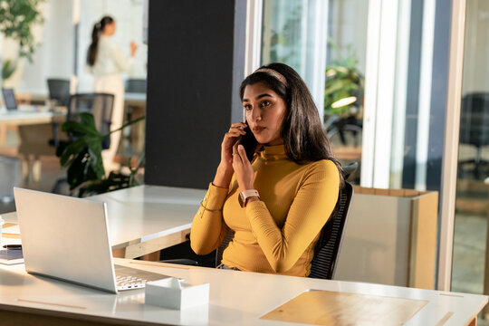 Indian female wearing turtleneck speaking on smartphone at office desk with laptop and tissue box