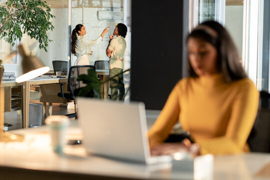 Diverse coworkers discussing diagrams on glass partition in open plan office with laptop