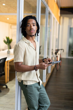 African American man leaning on glass partition in open office wearing linen shirt, holding tablet
