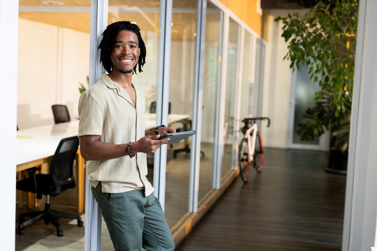 African American man leaning against glass partition holding tablet in office hall, copy space