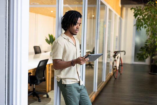 African American man in beige shirt leaning on glass wall in office corridor holding tablet