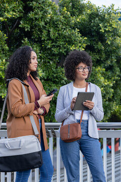 Diverse female friends standing by metal railing on park path holding smartphone, using tablet