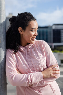 Woman leaning against metal pole and looking right on rooftop parking deck wearing pink jacket