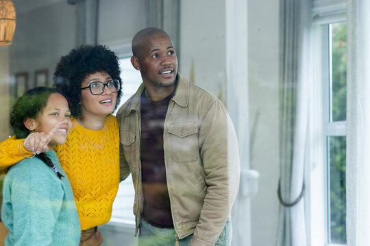 Diverse family looking out through large window in living room, framed by curtains and hanging lamp